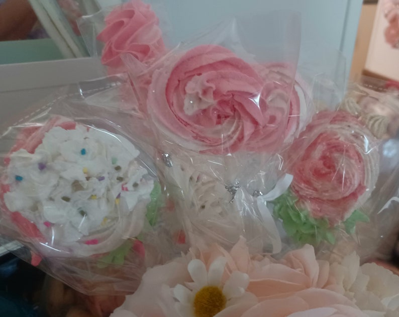 May include: Close-up of several pink and white meringue lollipops and cupcake-shaped treats, wrapped in clear cellophane. The lollipops are shaped like roses and swirls. The cupcake has white frosting with colorful sprinkles.