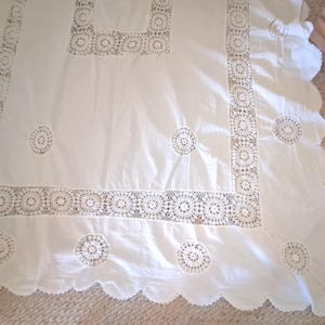 May include: A white rectangular tablecloth featuring intricate crochet lace borders and circular lace insets. The fabric has a smooth texture, and the edges are finished with a scalloped hem, creating a decorative pattern across the textile surface.