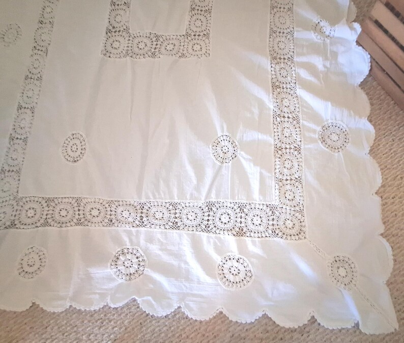 May include: A white rectangular tablecloth featuring intricate crochet lace borders and circular lace insets. The fabric has a smooth texture, and the edges are finished with a scalloped hem, creating a decorative pattern across the textile surface.