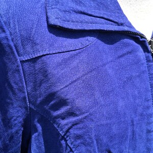 May include: A vibrant royal blue jacket featuring a textured suede-like finish, a classic pointed collar, and visible topstitching along the shoulder yoke and front panels. The garment includes a dark zipper closure extending up to the neckline.
