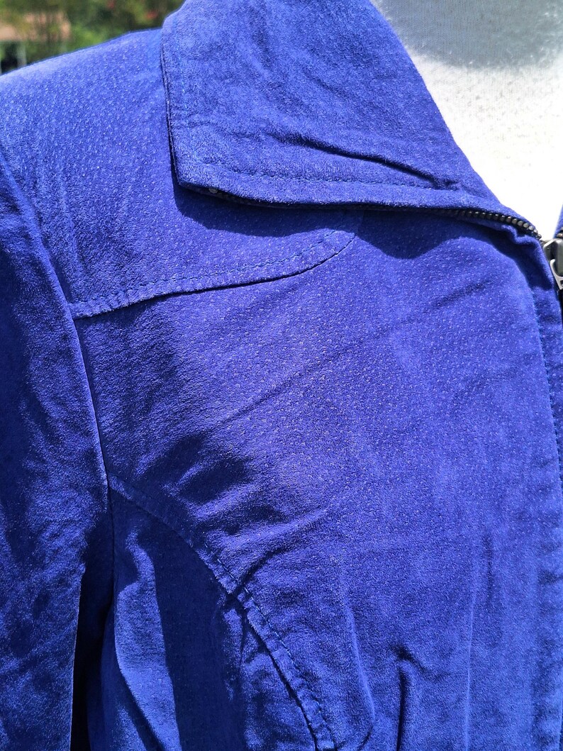 May include: A vibrant royal blue jacket featuring a textured suede-like finish, a classic pointed collar, and visible topstitching along the shoulder yoke and front panels. The garment includes a dark zipper closure extending up to the neckline.