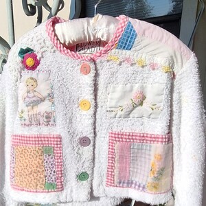 May include: A white terry cloth jacket featuring a pink gingham trim, four colorful buttons, and various fabric patches including a ballerina illustration, floral embroidery, and patterned squares. The label reads FLOWER LADY.