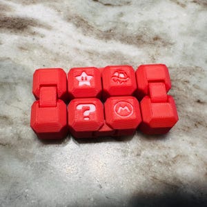 May include: A red, articulated fidget toy with eight connected cube-shaped blocks. Each block displays a white icon: a star, a question mark, a mushroom, and the letter "M". The toy is designed for manipulation and play.