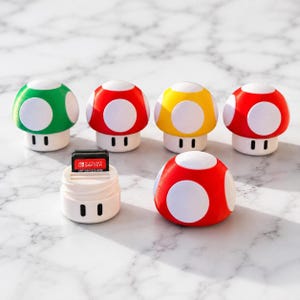 Op de afbeelding: Een verzameling van zes paddenstoelvormige opbergbakjes in groen, rood en geel met witte stippen. Eén bakje is geopend en toont een zwarte Nintendo Switch-spelcartridge. Elk bakje heeft een witte basis met twee zwarte verticale lijnen.