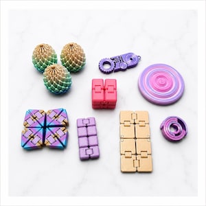 Puede incluir: Una colección de juguetes fidget coloridos en varias formas. Incluye tres juguetes en forma de huevo con degradados verdes, dorados y azules, un bloque rosa, un engranaje morado y un juguete dorado en forma de cadena. Otros juguetes son morados, rosas y azules.