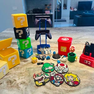 May include: A selection of Super Mario-themed items, including question mark blocks, character patches, controller stands, and a red storage box. The items are displayed on a marble surface, showcasing a variety of gaming accessories and collectibles.