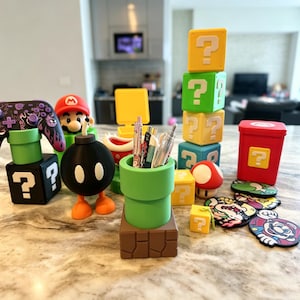 May include: A collection of Super Mario-themed desk accessories, including a green pipe pen holder, question mark blocks, a Bob-omb figure, and a red mushroom. The items are in various colors, including yellow, green, red, and black.