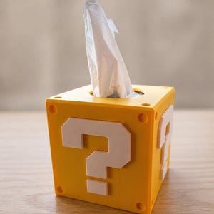 Könnte beinhalten: Gelbe Taschentuchbox in Form eines Fragezeichenblocks aus einem Videospiel. Die Box hat weiße Fragezeichen auf zwei Seiten und eine runde Öffnung oben mit weißen Taschentüchern, die herausragen. Die Box hat eine quadratische Form mit abgerundeten Ecken.