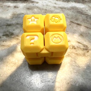 May include: A yellow cube-shaped toy with symbols from a popular video game series. The symbols include a star, a question mark, a mushroom, and a flower. The toy is made of a solid, matte material.