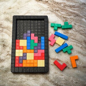 May include: A black Tetris game board with colorful, interlocking game pieces. The pieces are various shapes and colors, including red, blue, green, yellow, and pink. The word "TETRIS" is printed on the bottom of the board.