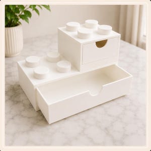 以下が含まれることがあります： 2段構造のブロック型をした白いプラスチック製のデスクオーガナイザー。下段には幅の広い引き出しがあり、上段には小さな引き出しと、おもちゃのブロックを模した4つの円形の突起が表面に付いています。
