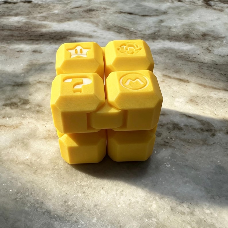 May include: A yellow cube toy featuring embossed symbols such as a crown, question mark, and flower. The toy, made from a solid, matte material, could be a puzzle or building block set. It sits on a light-coloured, textured surface.