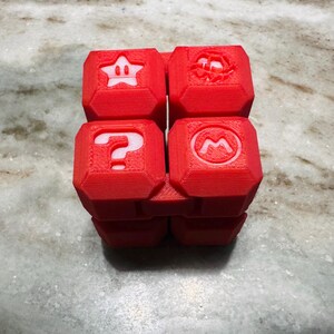 May include: A red, 3D-printed puzzle cube with symbols from the Super Mario video game series. The cube shows a star, a question mark, a mushroom, and a flower. The cube is on a marble surface.