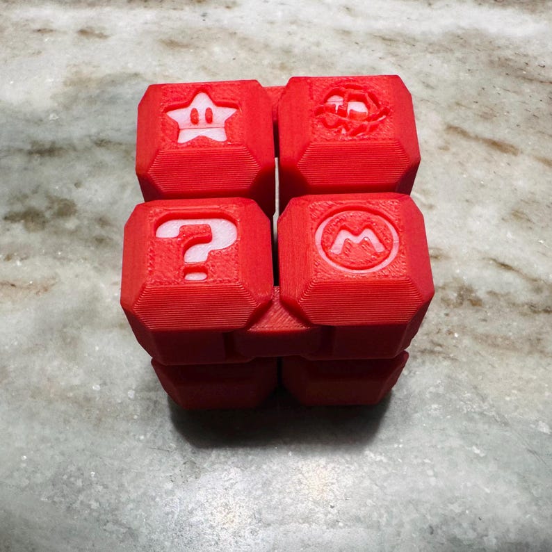 May include: A red, 3D-printed puzzle cube with symbols from the Super Mario video game series. The cube shows a star, a question mark, a mushroom, and a flower. The cube is on a marble surface.