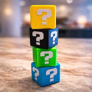May include: Four brightly coloured question mark blocks stacked on top of each other. The top block is yellow, followed by black, green, and blue. Each block features a white question mark. The blocks are on a wooden surface.