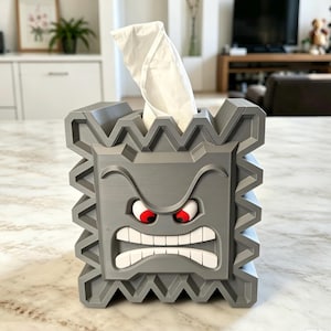 Puede incluir: Una funda para caja de pañuelos gris de forma cuadrada que representa a un personaje Thwomp. Presenta un borde dentado y puntiagudo, una expresión facial severa con cejas fruncidas, ojos rojos y una boca ancha que muestra dientes blancos.