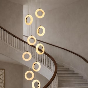 May include: A contemporary chandelier featuring multiple circular lights suspended from the ceiling. Each light has a gold-coloured ring with a glowing, warm-toned centre. The lights are arranged in a vertical cascade, illuminating a staircase.