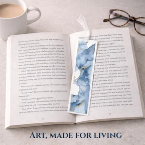 Puede incluir: Un marcapáginas de acuarela en tonos azules y dorados con una cinta blanca, colocado en un libro abierto. El marcapáginas tiene un diseño abstracto. El texto "ART, MADE FOR LIVING" está en la parte inferior.