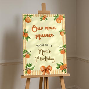 Op de afbeelding: Een aquarel-stijl bord op een ezel, met de tekst "Our main squeeze" en "Welcome to Mira's 1st birthday." Het bord is versierd met sinaasappel illustraties en een strik. De ezel is van hout.