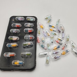 Może przedstawiać: Czarny smartfon z dwunastoma przezroczystymi plastikowymi kapsułkami przymocowanymi do powierzchni, z których każda zawiera maleńką, kolorową figurkę. Obok telefonu znajduje się zbiór licznych, identycznych przezroczystych kapsułek z figurkami.