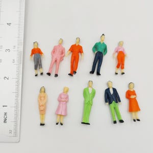 May include: A collection of miniature figurines depicting people in various outfits. The figures are painted in bright colors, including orange, pink, green, and blue. Each figure is approximately 1 inch tall, perfect for model train sets or architectural models.