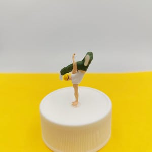 Puede incluir: Una figura en miniatura de un breakdancer en posición de handstand. La figura lleva pantalones cortos blancos, un casco amarillo y una chaqueta verde. Está sobre una superficie blanca con un fondo amarillo y blanco.