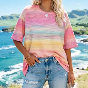 May include: A short-sleeved, crew neck t-shirt with a horizontal tie-dye pattern in shades of pink, yellow, and blue. The shirt is paired with light blue, distressed jeans. The model is wearing a silver necklace and bracelets.