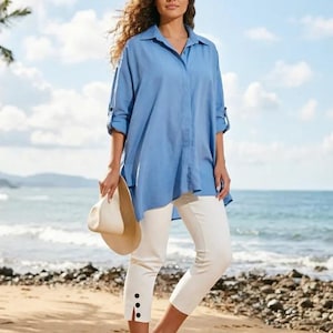 May include: A woman wearing a light blue button-down shirt and white capri pants stands on a beach. The shirt has rolled-up sleeves. She holds a beige sun hat. The pants have black buttons at the ankle. Gold sandals complete the outfit.