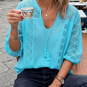 May include: A light blue blouse with intricate lace detailing on the sleeves and upper chest. The blouse has a V-neck and three-quarter sleeves. The person is wearing multiple gold necklaces and bracelets, and a watch with a brown leather strap. The person is holding a small cup of coffee.