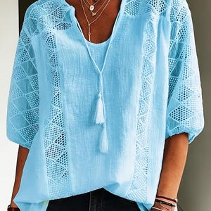 May include: Light blue blouse with a V-neck and lace detailing on the sleeves and sides. The blouse has a tassel tie and is paired with black jeans. A layered necklace is visible.