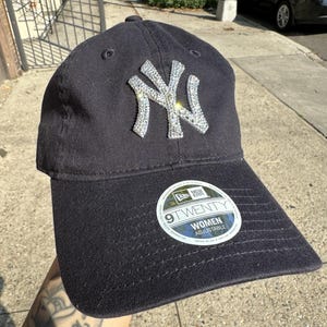 May include: A black baseball cap featuring a prominent New York Yankees logo embellished with clear rhinestones on the front. The cap includes a circular sticker on the brim with the text New Era 9TWENTY WOMEN ADJUSTABLE in white and black.