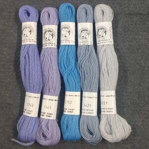 May include: Five skeins of embroidery floss in shades of lavender, blue, and gray. The skeins are labeled with the numbers 1342, 1341, 2152, 1421, and 3221. The labels also indicate that the floss is "Pure Laine/Wool" and "Colour Fast and Teint".