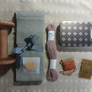 May include: A wooden spool, a gray fabric wrap tied with a blue ribbon, a skein of pink thread, a patterned tin, a spool of orange thread, and a small piece of tan fabric with pins. The items are arranged on a light-colored surface.