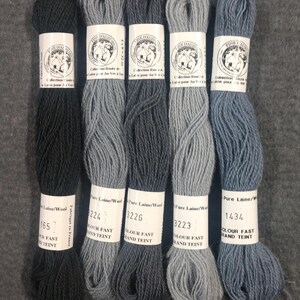 May include: Five skeins of wool yarn in shades of gray and blue, arranged vertically. Each skein has a paper label with text and a logo. The yarn is labeled as "Pure Laine/Wool" and "Colour Fast Grand Teint".