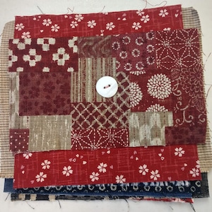 May include: A patchwork quilt block made of various red and beige fabrics with a white button in the center. The block is made up of squares and rectangles of different sizes and patterns.