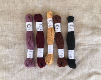Può includere: Cinque matasse di filo da ricamo in lana disposte verticalmente. I colori da sinistra a destra sono malva, bordeaux intenso, giallo dorato, marrone scuro e nero. Ogni matassa presenta un'etichetta di carta bianca con testo e un logo.