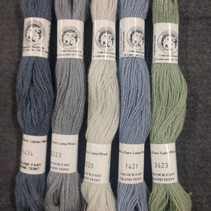 May include: Five skeins of wool yarn in different colors: blue, gray, white, light blue, and green. Each skein has a label with the brand name "Fine Dauphine", the collection name "Collection Route", and the color number. The labels also say "Pure Laine/Wool" and "Colour Fast Grand Teint".
