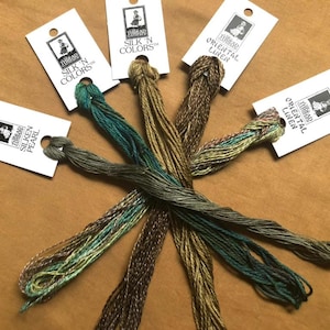 May include: Assortment of embroidery threads in various colors, including green, brown, and gold. Each thread bundle is attached to a white tag with a logo and product information. The threads are arranged on a brown surface, creating a visually appealing display.
