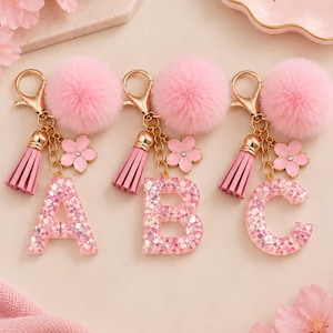 May include: Three keychains featuring the letters A, B, and C in clear resin filled with pink glitter. Each keychain includes a gold-tone lobster clasp, a fluffy pink pom-pom, a pink faux leather tassel, and a small pink flower charm with a center crystal.