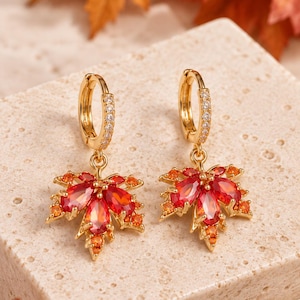 May include: A pair of gold hoop earrings featuring a dangling maple leaf charm. Each leaf is encrusted with vibrant red pear-shaped stones and small orange round stones. The hoop portion of the earring is decorated with a row of clear white stones.