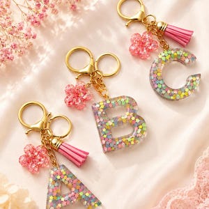 May include: Three letter keychains featuring the letters A, B, and C. Each keychain is made of clear resin filled with small, multi-coloured star-shaped glitter. They include a gold-tone metal ring, a pink flower charm, and a pink tassel.