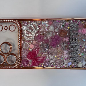 Pu&ograve; includere: Una custodia per telefono color oro rosa con un centro trasparente pieno di abbellimenti rosa e trasparenti. La custodia presenta una variet&agrave; di forme, tra cui fiori, fiocchi e cristalli. La parola "JENNA" &egrave; scritta in lettere argentate.