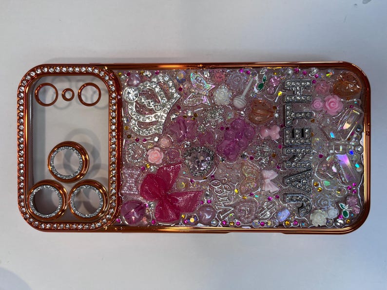 Pu&ograve; includere: Una custodia per telefono color oro rosa con un centro trasparente pieno di abbellimenti rosa e trasparenti. La custodia presenta una variet&agrave; di forme, tra cui fiori, fiocchi e cristalli. La parola "JENNA" &egrave; scritta in lettere argentate.