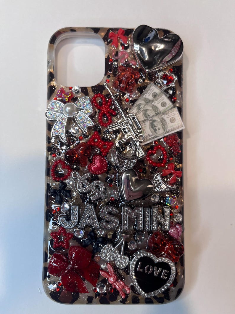 Pu&ograve; includere: Custodia decorativa per telefono con sfondo a stampa leopardata. Gli abbellimenti includono strass, fiocchi, cuori, una pistola giocattolo e una piccola pila di banconote. Il nome "JASMIN" &egrave; scritto con strass e un ciondolo a forma di cuore recita "LOVE".