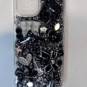 Pu&ograve; includere: Custodia trasparente per telefono decorata con abbellimenti neri e argento. Include una stella, un cuore, una croce, un fiocco e una chitarra. Sono visibili anche piccoli mucchi di banconote da 100 dollari. La custodia &egrave; ricoperta di glitter e piccole perle d'argento.