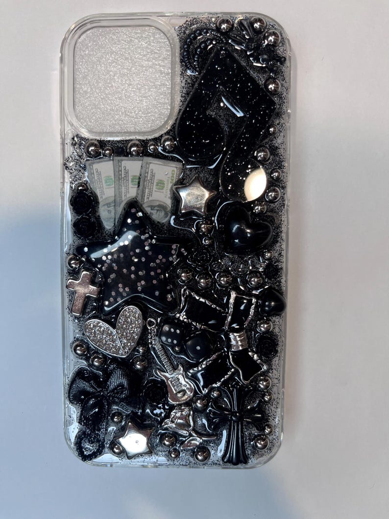 Pu&ograve; includere: Custodia trasparente per telefono decorata con abbellimenti neri e argento. Include una stella, un cuore, una croce, un fiocco e una chitarra. Sono visibili anche piccoli mucchi di banconote da 100 dollari. La custodia &egrave; ricoperta di glitter e piccole perle d'argento.