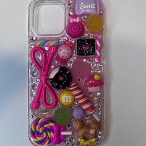 Pu&ograve; includere: Una custodia trasparente per telefono decorata con varie caramelle colorate e strass. La custodia presenta un fiocco rosa, un lecca-lecca, un cupcake e una barretta di caramelle con la scritta "Sweet".