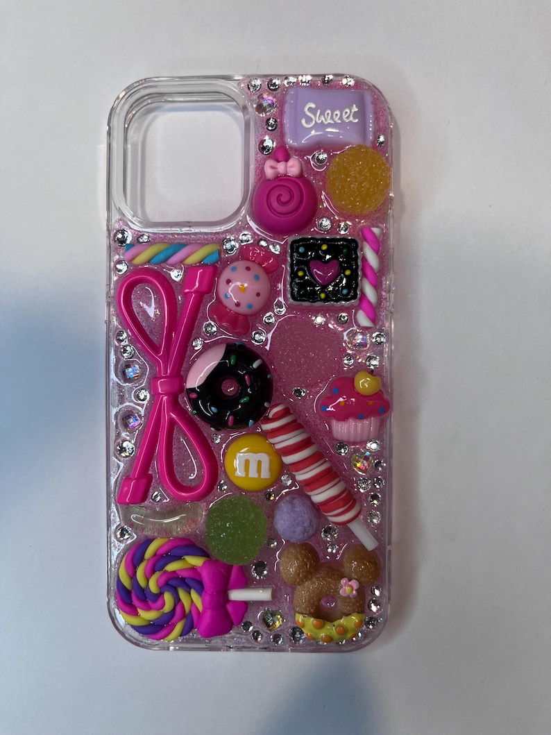 Pu&ograve; includere: Una custodia trasparente per telefono decorata con varie caramelle colorate e strass. La custodia presenta un fiocco rosa, un lecca-lecca, un cupcake e una barretta di caramelle con la scritta "Sweet".
