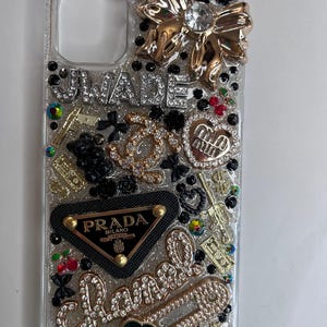 Pu&ograve; includere: Custodia trasparente per telefono impreziosita da fiocco dorato, strass e vari ciondoli. Presenta i testi "UWADE" e "Chanel" in strass, insieme a un logo Prada e altri elementi decorativi. La custodia &egrave; ricoperta di glitter.