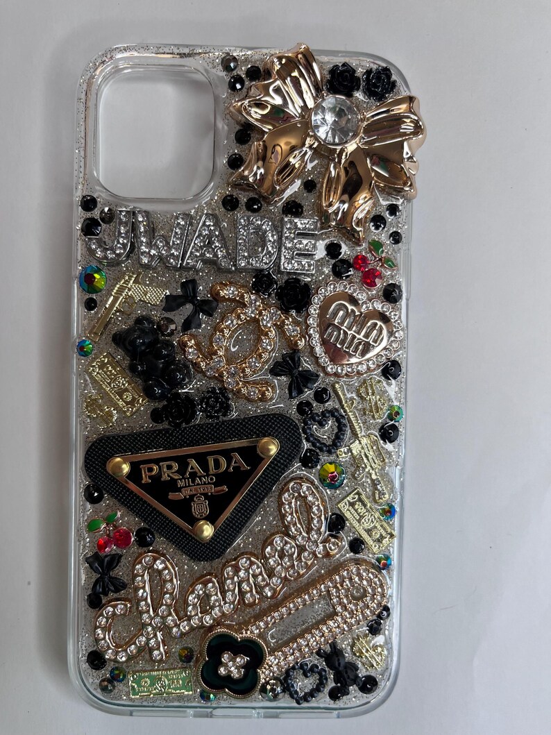 Pu&ograve; includere: Custodia trasparente per telefono impreziosita da fiocco dorato, strass e vari ciondoli. Presenta i testi "UWADE" e "Chanel" in strass, insieme a un logo Prada e altri elementi decorativi. La custodia &egrave; ricoperta di glitter.